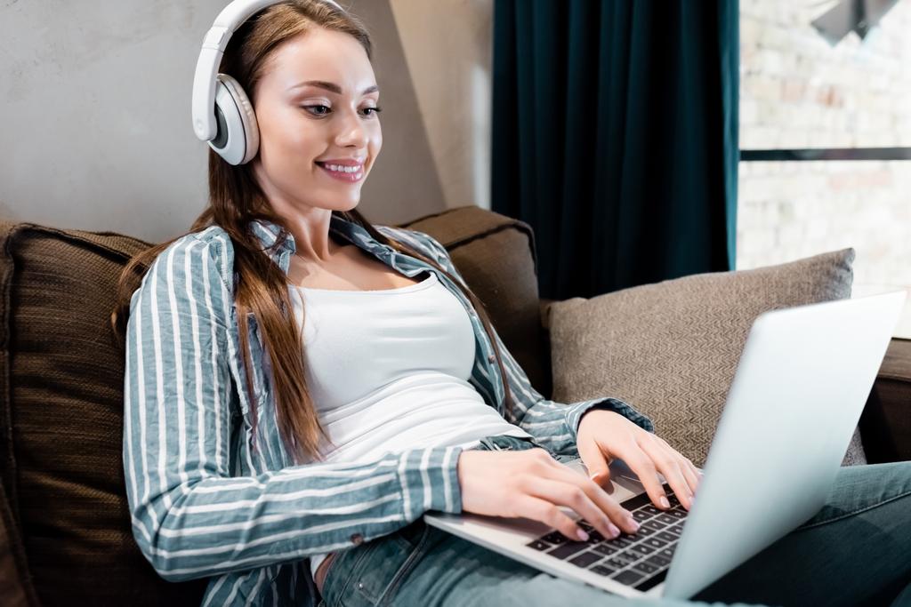 επιλεκτική εστίαση του χαρούμενος ελεύθερος επαγγελματίας σε ασύρματα ακουστικά χρησιμοποιώντας φορητό υπολογιστή στο σαλόνι  - Φωτογραφία, εικόνα