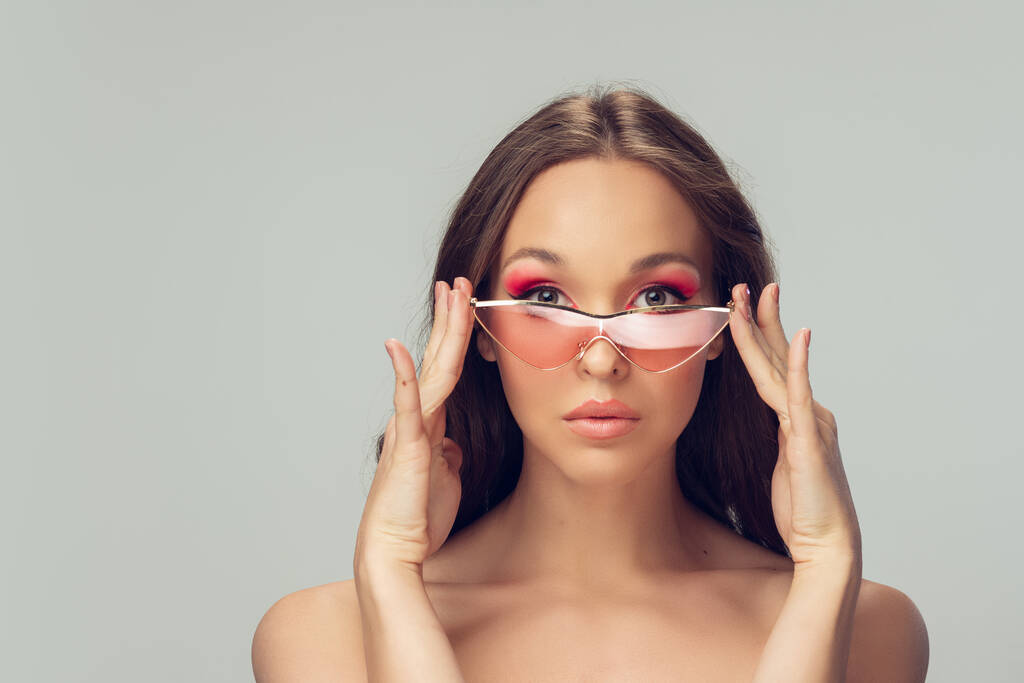 Primer plano de la hermosa mujer joven con el pelo rizado largo y saludable y maquillaje brillante usando gafas de color rosa con estilo aislado en el fondo del estudio gris
 - Foto, Imagen
