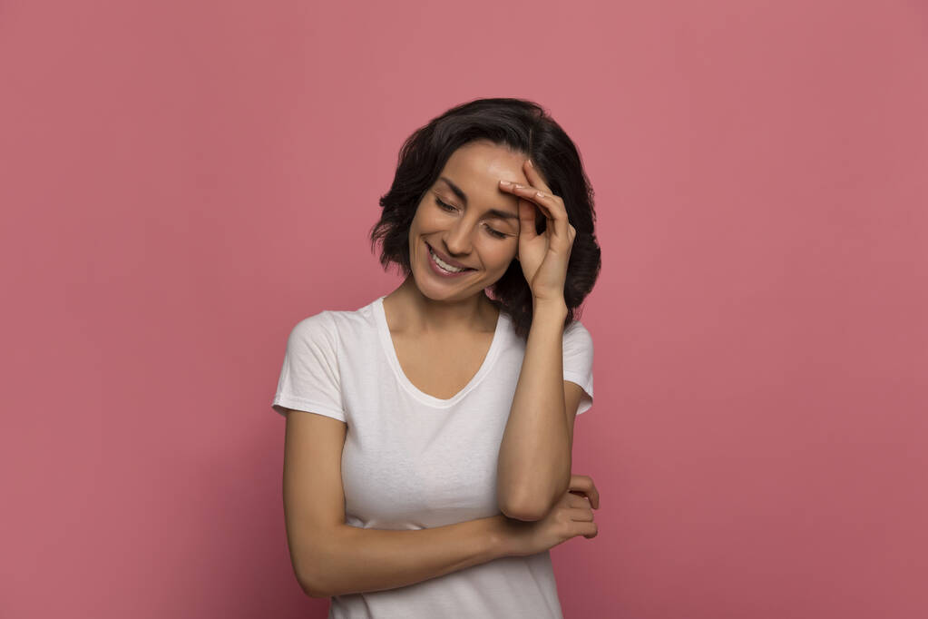 Schönheitsgeheimnisse. Nahaufnahme einer modernen jungen Frau in weißem T-Shirt, die mit geschlossenen Augen lächelt, während sie mit der linken Hand ihre Stirn berührt. - Foto, Bild