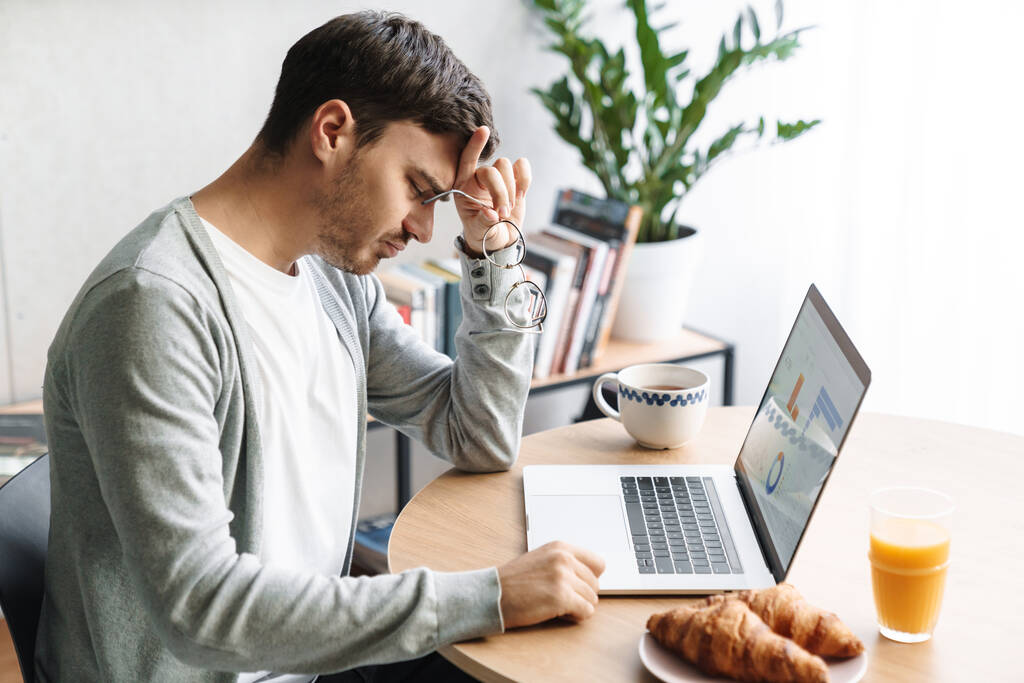 Beeld van vermoeide jongeman met bril die vermoeidheid uitdrukt tijdens het werken aan laptop thuis - Foto, afbeelding