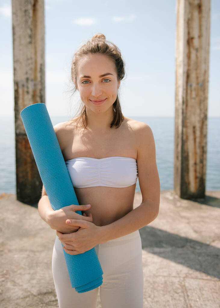 Улыбающаяся молодая женщина, держащая коврик для йоги, стоя на пирсе у моря
 - Фото, изображение