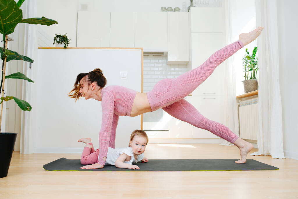 Una giovane madre in abbigliamento sportivo pratica ginnastica a casa, in piedi nel bar alza alternativamente le gambe, accanto a striscia giovane figlia
 - Foto, immagini