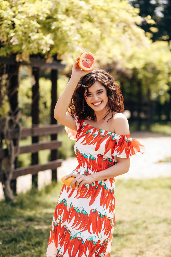 Mode Outdoor-Foto von schönen sinnlichen Frau mit dunklen Haaren in luxuriösen Kleid mit Accessoires, hält Grapefruit, posiert im Sommergarten - Foto, Bild