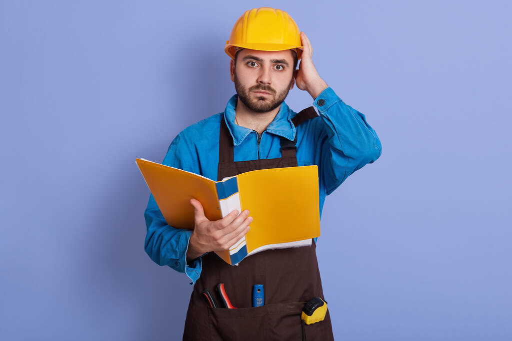 Portrait of emotional magnetic helpless young builder looking directly at camera, holding papercase, putting one hand on helmet, having working day, being busy. People and work problems concept. - Photo, Image
