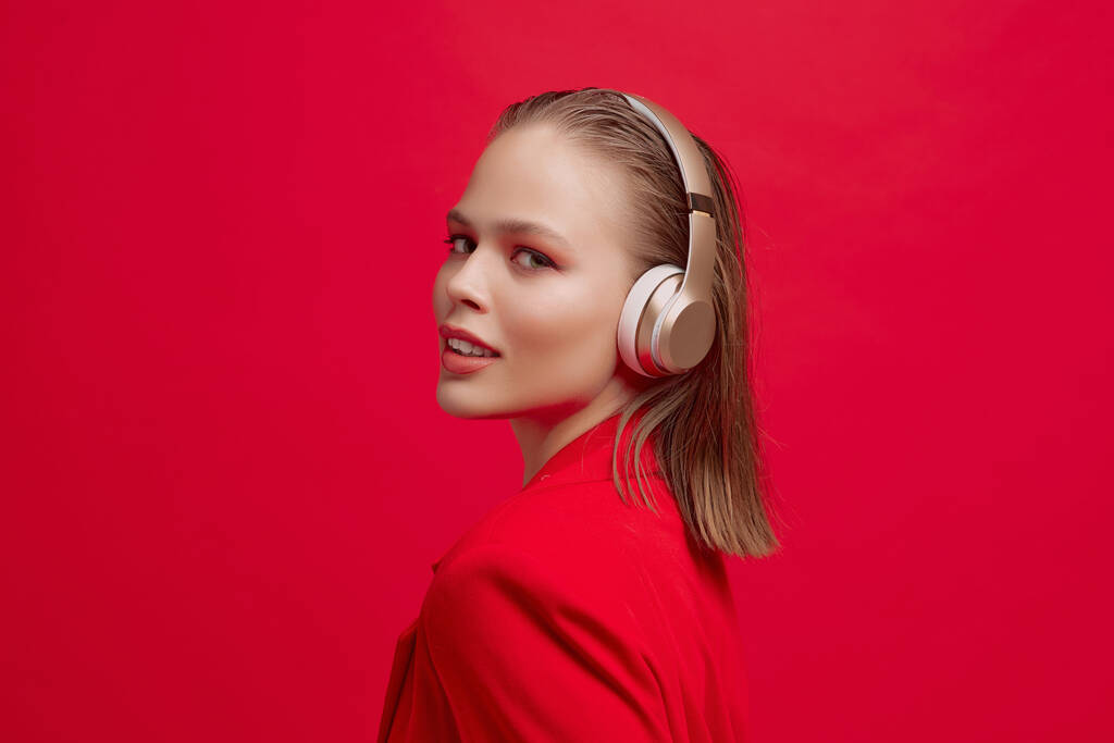 Uma menina bonita com um belo penteado está ouvindo música em fones de ouvido sem fio em um fundo vermelho
. - Foto, Imagem