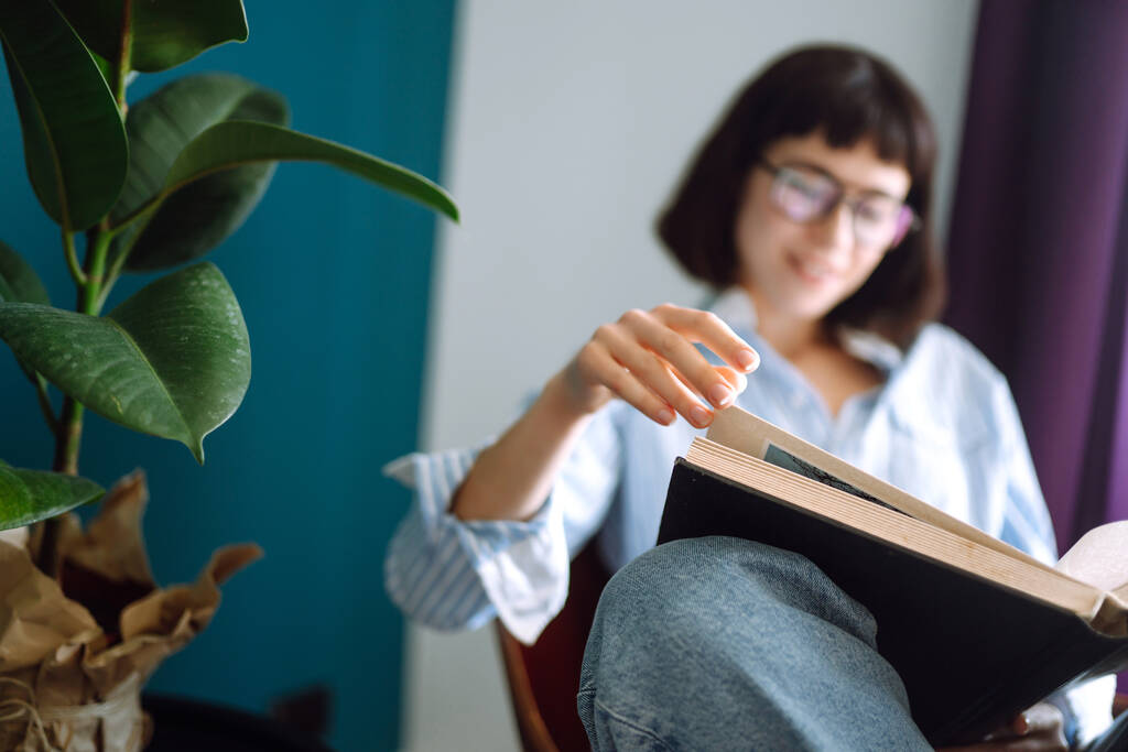 Nuori nainen lukee kirjaa kotona, istuu modernilla tuolilla rentoutuu olohuoneessaan. Käsitys levosta, rentoutumisesta, mukavuudesta
.  - Valokuva, kuva