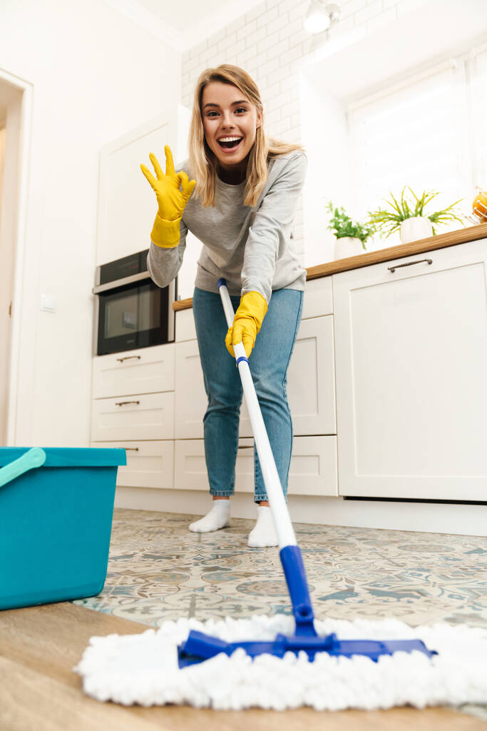 Foto de alegre joven ama de casa con guantes haciendo gestos signo de ok mientras fregaba piso en la cocina moderna
 - Foto, imagen