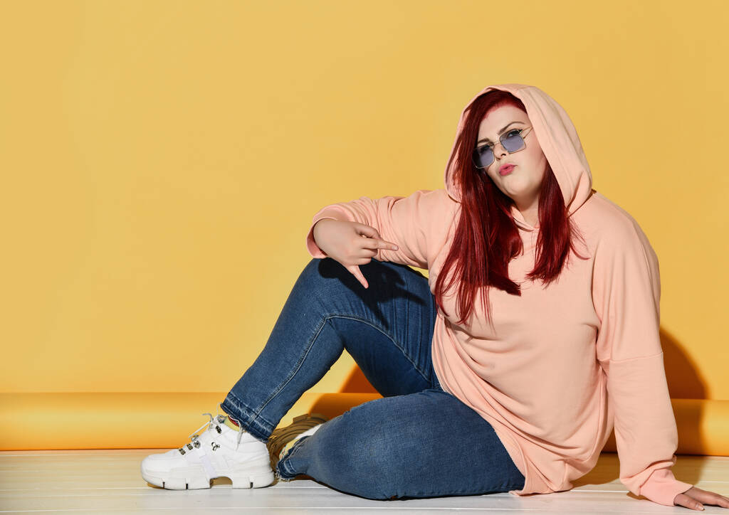 Jovem sorrindo mulher com excesso de peso de cabelos vermelhos em roupas casuais, tênis e óculos de sol sentados no chão e se sentindo legal
 - Foto, Imagem