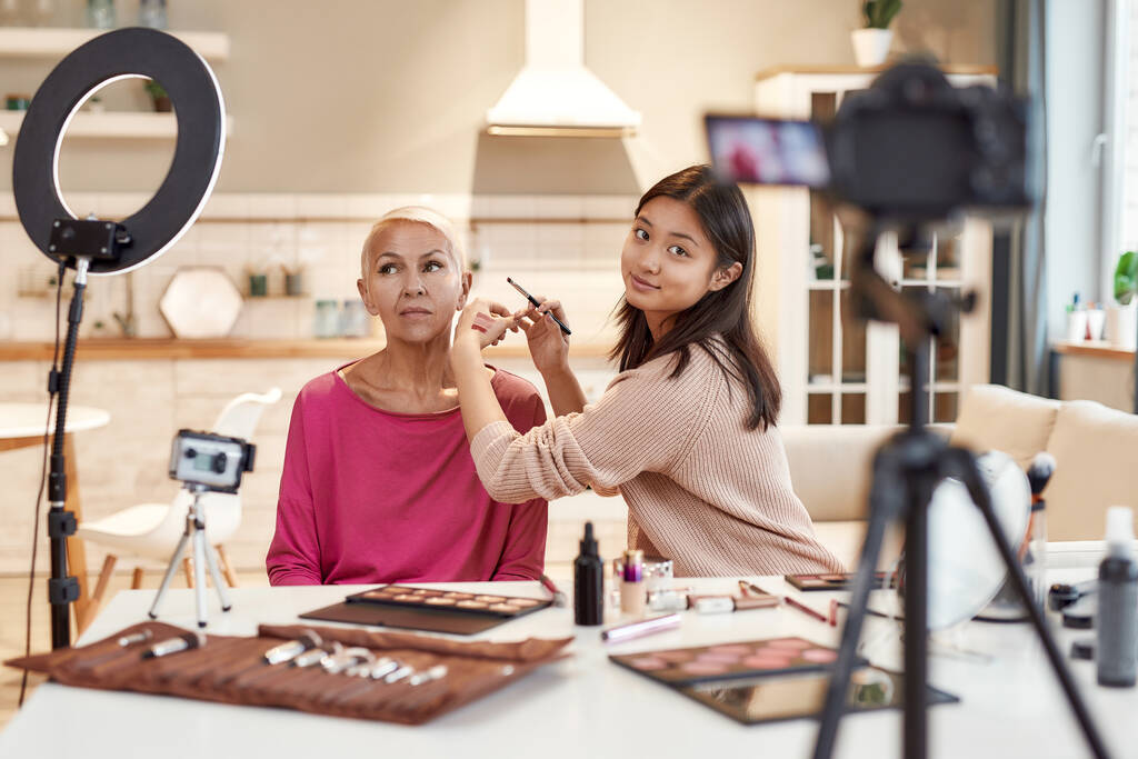 Être glamour. Blogueuse asiatique montrant les couleurs du rouge à lèvres tout en appliquant le maquillage sur le visage de la femme d'âge moyen en utilisant des cosmétiques sur la table. Deux femmes enregistrant une vidéo de tutoriel pour le blog de beauté
 - Photo, image