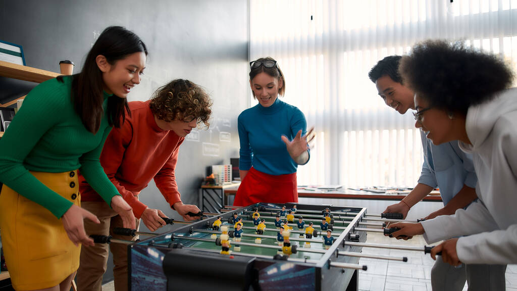 Обеденный перерыв. Группа молодых и счастливых мультикультурных людей в повседневной одежде играет в настольный футбол в современном офисе и смеется. Деятельность офиса. Веселитесь вместе. Счастливые сотрудники
 - Фото, изображение