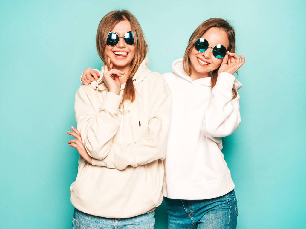 Две молодые красивые блондинки улыбающиеся хипстеры в модной летней одежде с капюшоном. Сексуальные беззаботные женщины позируют у голубой стены. Модные и позитивные модели, веселые в солнечных очках - Фото, изображение