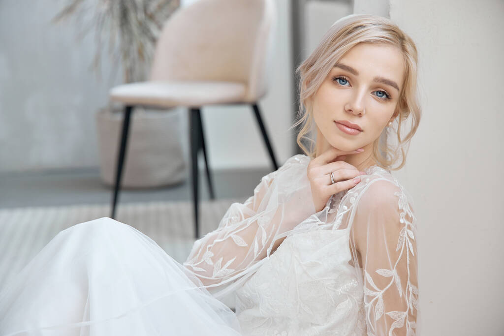 Ideal bride sitting on the floor, portrait of a girl in a long white dress. Beautiful hair and clean, soft skin. Wedding hairstyle blonde - Photo, Image