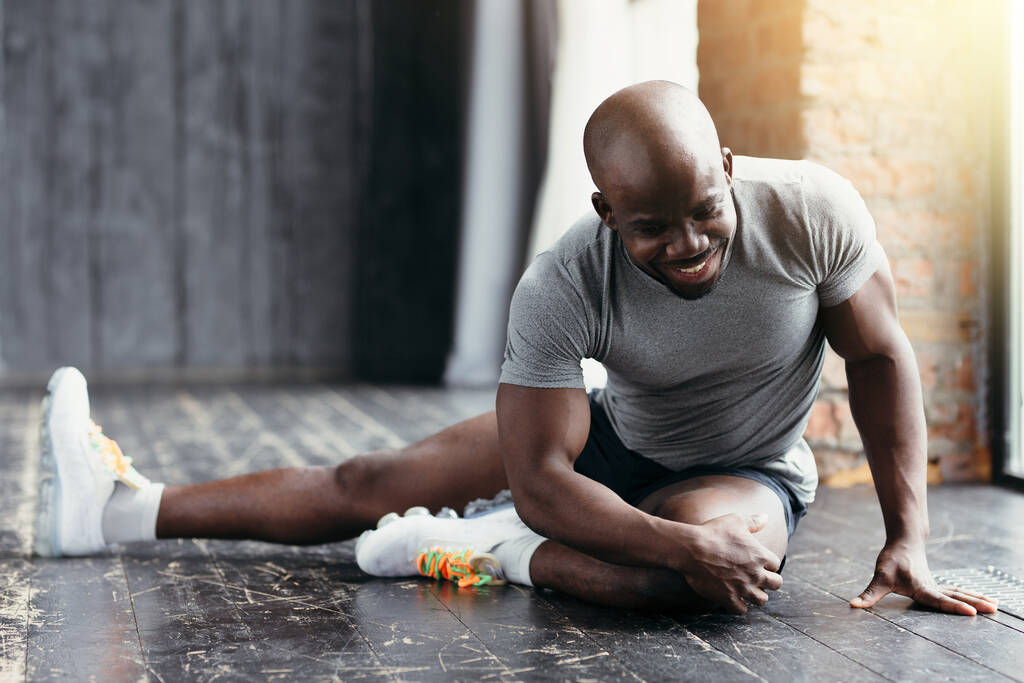 Een donkergekleurde glimlachende fitnesstrainer in een grijs T-shirt zit op de grond en doet oefeningen aan de zijkant. - Foto, afbeelding