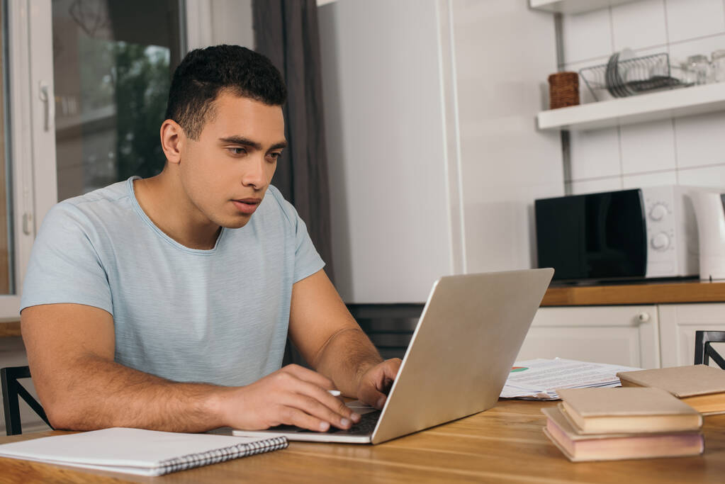 handsome mixed race freelancer using laptop near books and notebook - Photo, Image