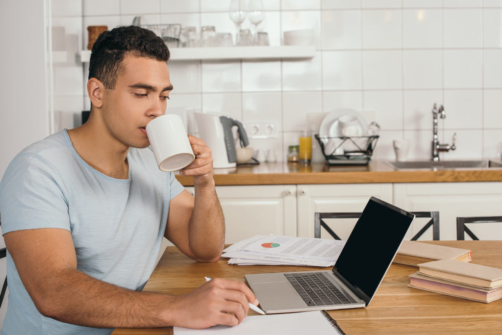 beau mixte homme de race tenant stylo et boire du café près d'un ordinateur portable avec écran blanc
 - Photo, image
