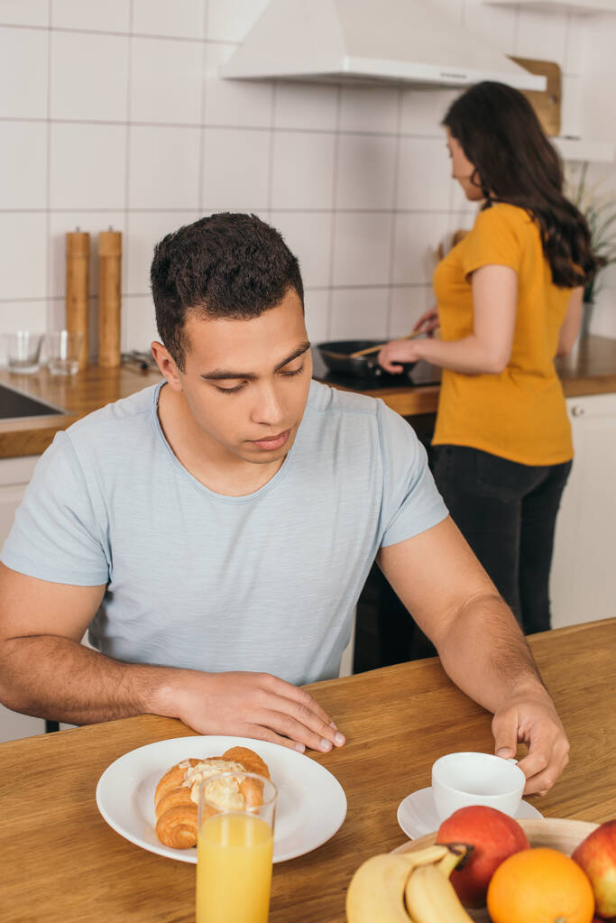 selective focus of handsome mixed race man touching cup near tasty croissant, fruits and orange juice on table  - Photo, Image