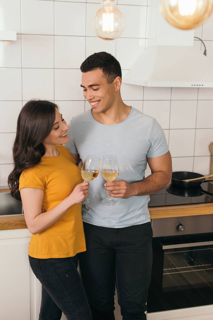 enfoque selectivo de feliz pareja interracial tintineo vasos de vino blanco en casa
 - Foto, Imagen
