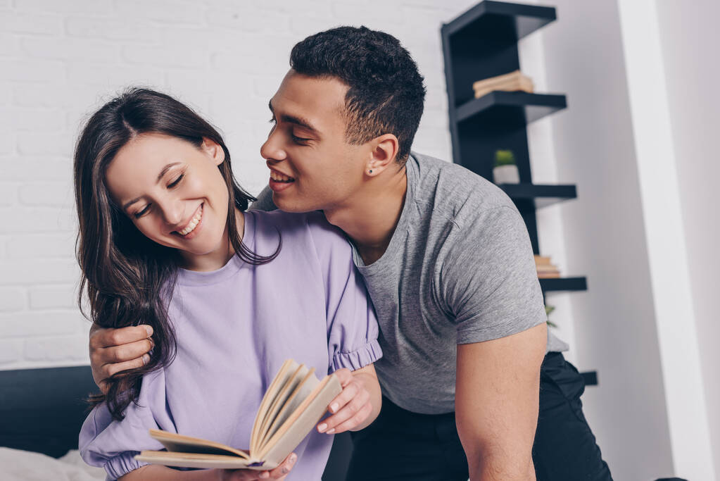 handsome mixed race man looking at smiling girl reading book - Photo, Image