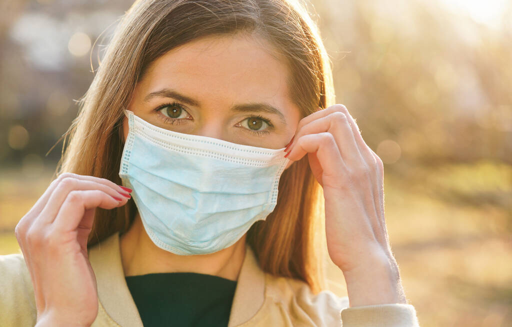 Young woman wearing blue disposable virus mouth nose mask, touching it with hands, nice sunny bokeh in background, closeup face portrait - Photo, Image