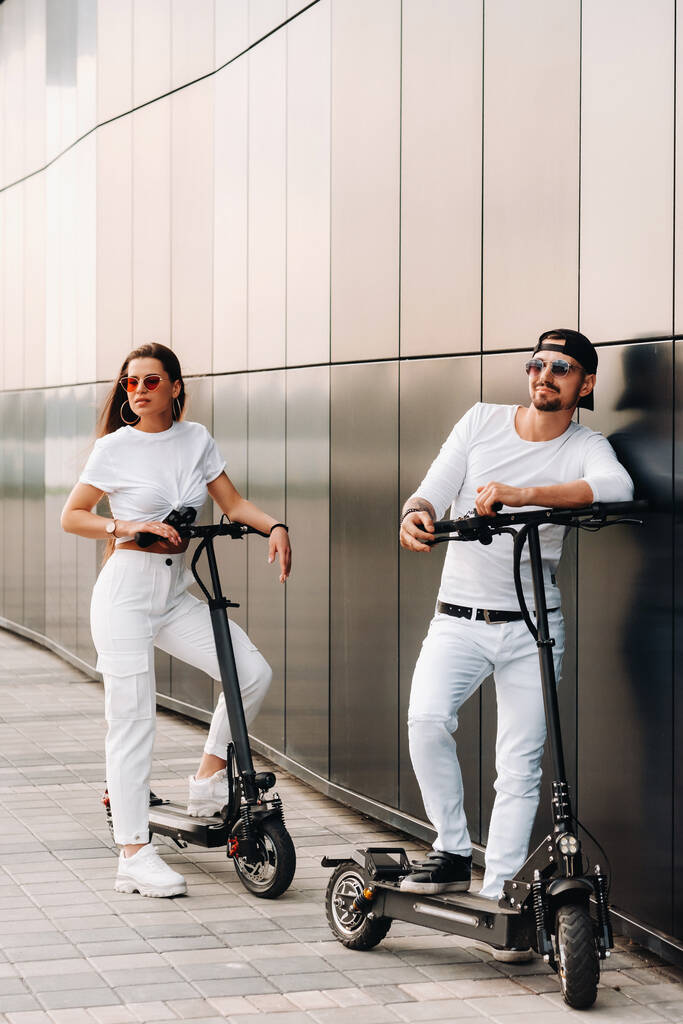 Bir kız ve bir adam şehirde elektrikli scooterlarla dolaşıyorlar. Birbirine aşık bir çift scooterlı. - Fotoğraf, Görsel