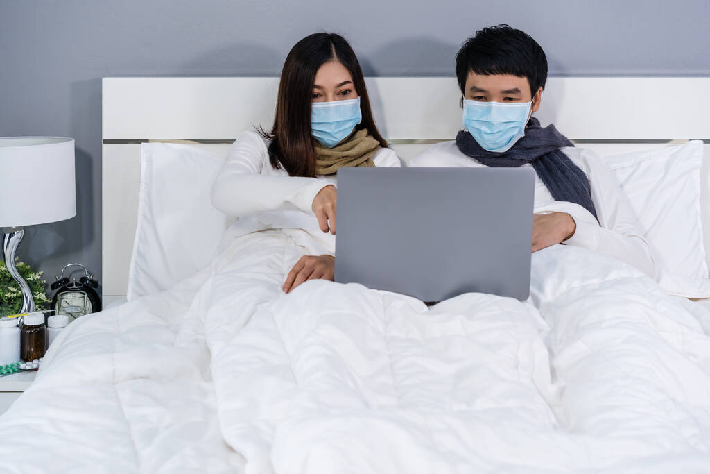 couple avec des masques médicaux à l'aide d'un ordinateur portable sur un lit pendant la quarantaine pandémique coronavirus (covid-19) rester à la maison. - Photo, image
