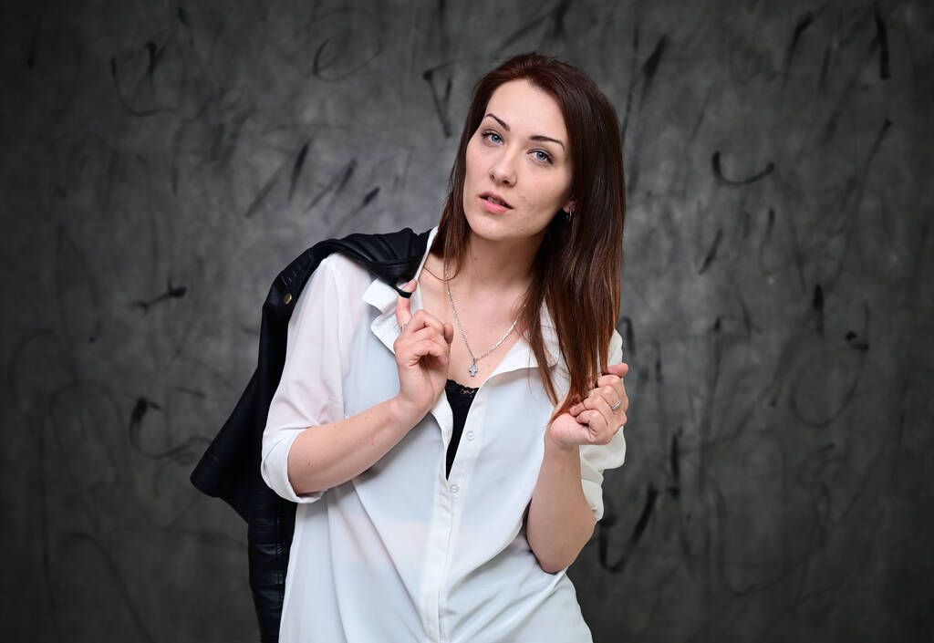 Ένα όμορφο μοντέλο στέκεται πάνω σε ένα γκρι φόντο με ένα λευκό πουκάμισο που κρατά ένα μαύρο σακάκι. Studio πορτρέτο ενός καυκάσιου μελαχρινού κοριτσιού με minimal μακιγιάζ. - Φωτογραφία, εικόνα