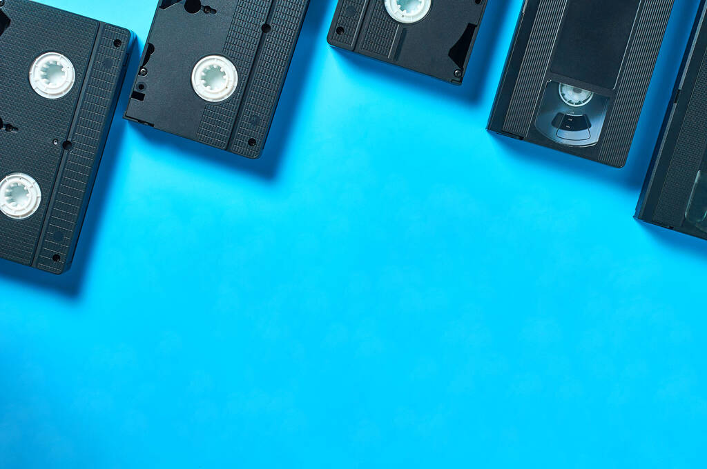 Ряд многих черных старых пластиковых VHS видеокассет рядом с пустым блокнотом лежит на синем столе. Концепция 90-х. Принято. Вид сверху
 - Фото, изображение