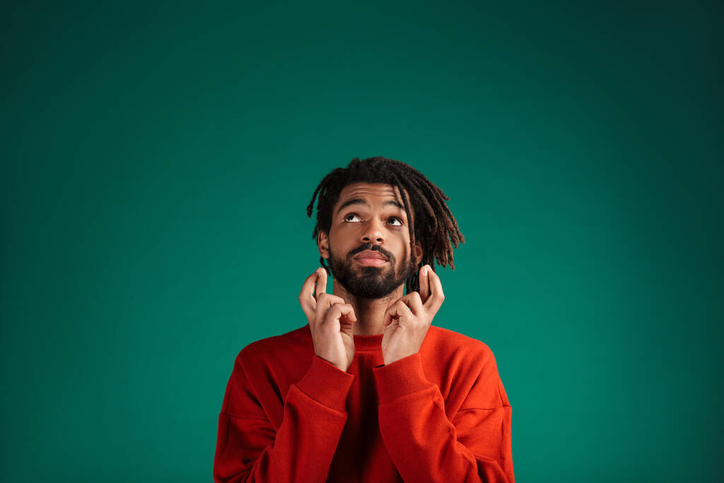 Portrait d'un jeune homme africain portant un pull debout isolé sur fond vert, les doigts croisés pour la bonne chance
 - Photo, image