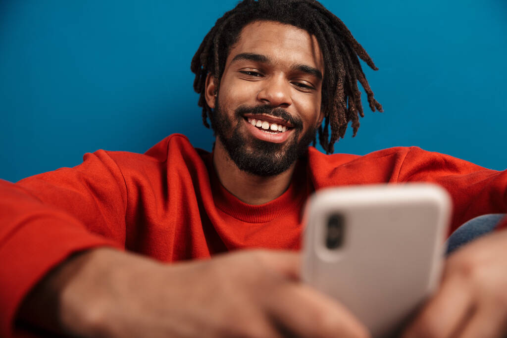 Close up portrait of a smiling young african man wearing pullover sitting isolated over blue background, using mobile phone - Photo, Image