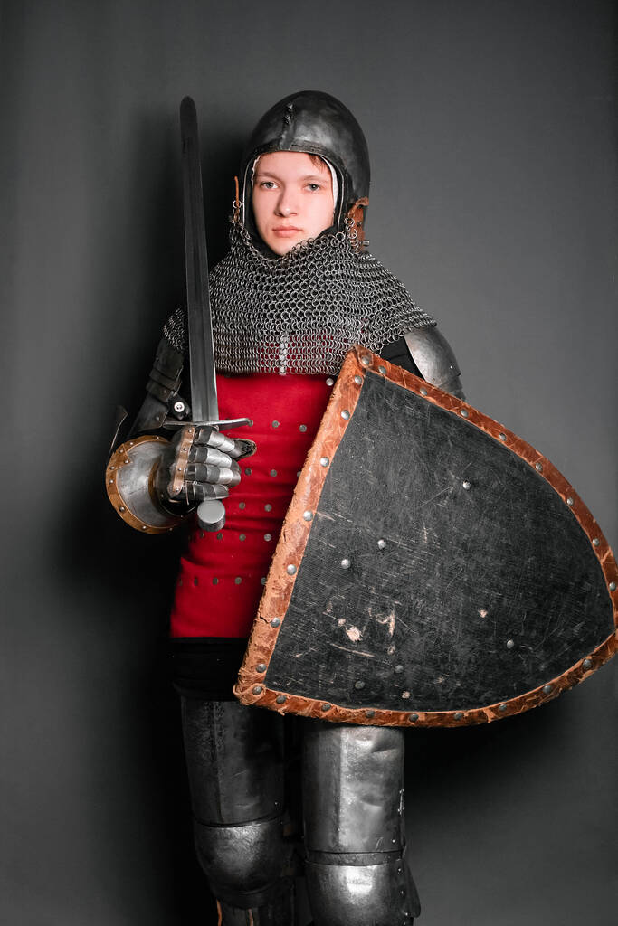 Młody rycerz w średniowiecznej zbroi uzbrojony w miecz i tarczę stojący na szarym tle. - Zdjęcie, obraz