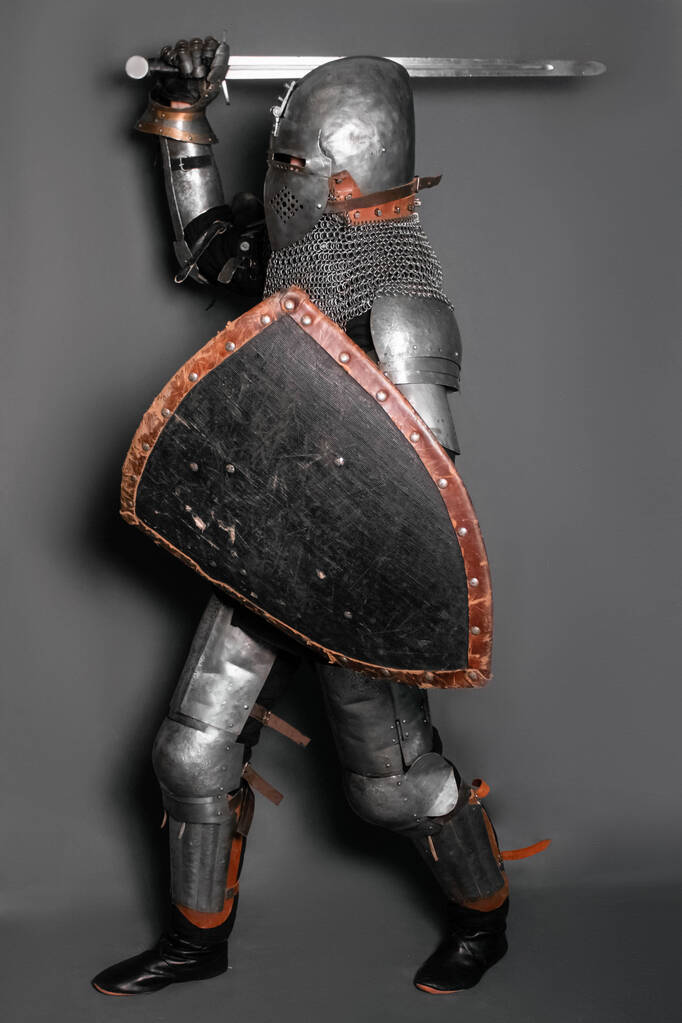 Un caballero medieval en armadura con un escudo y una espada en sus manos está en una posición de lucha y agita su arma
. - Foto, imagen