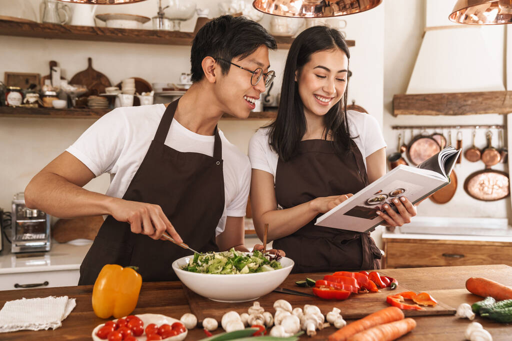 Imagen de pareja multicultural sonriendo y leyendo libro con recetas mientras se cocina la cena en acogedora cocina
 - Foto, imagen