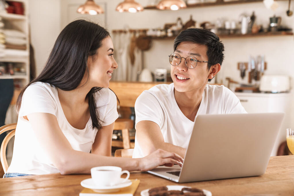 Kép a multikulturális boldog pár mosolyog és használja laptop, miközben ül az asztalnál a hangulatos konyhában - Fotó, kép