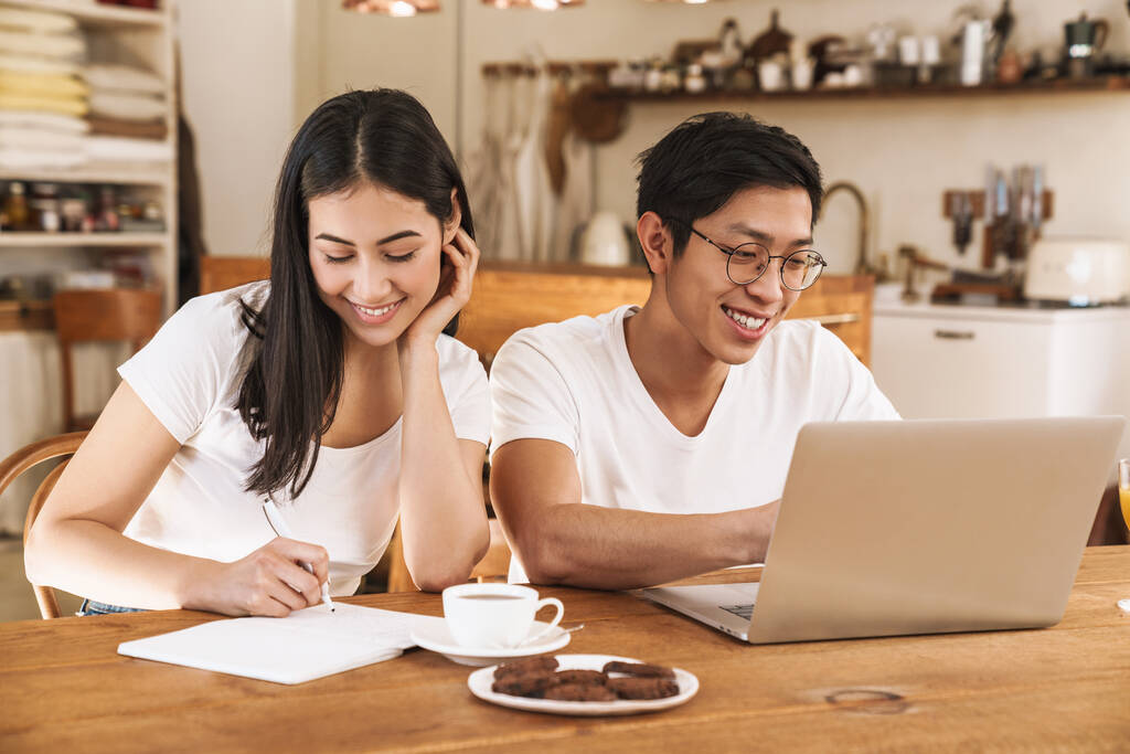 Kép mosolygó multikulturális pár jegyzetel a tervező és a laptop kényelmes konyhában - Fotó, kép