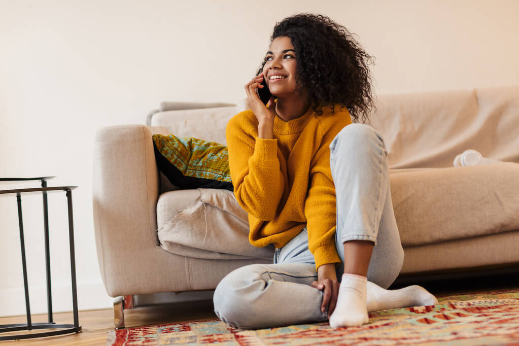 Imagen de la sonriente mujer afroamericana hablando por celular mientras está sentada en el suelo en la sala de estar
 - Foto, Imagen