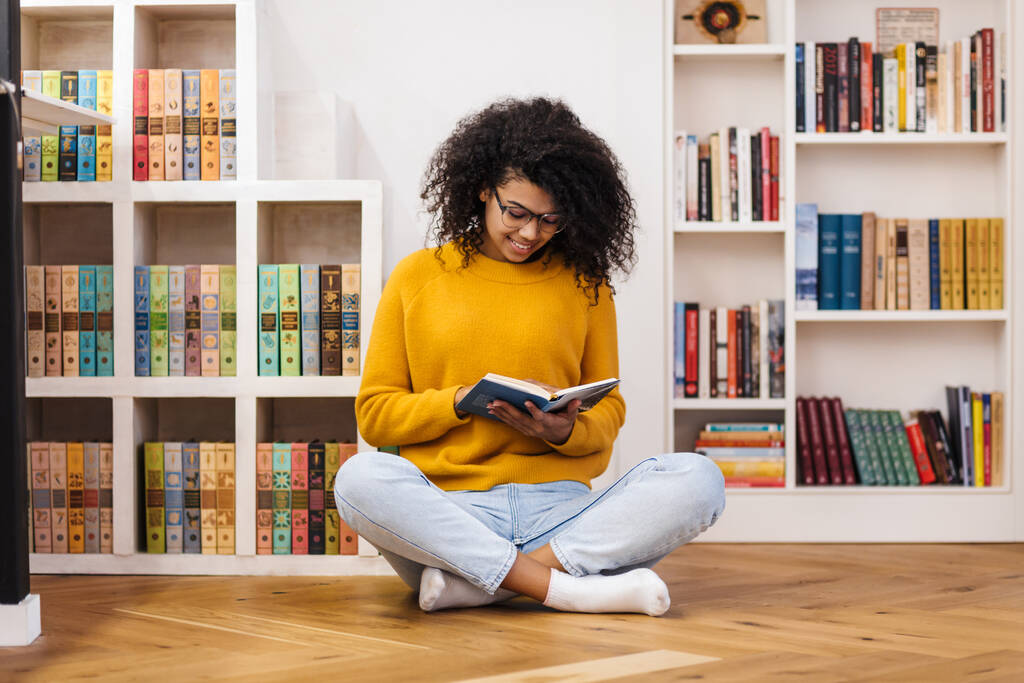 Image d'une femme afro-américaine joyeuse dans un livre de lecture de lunettes alors qu'elle était assise sur le sol dans le salon
 - Photo, image