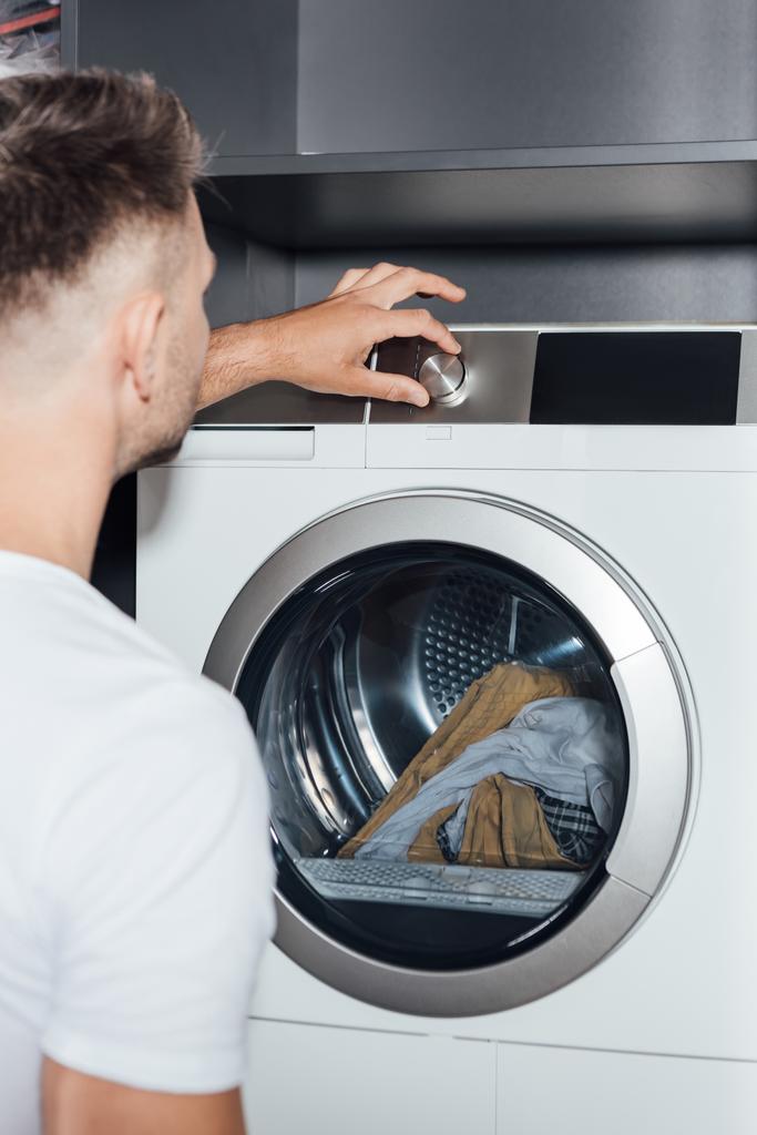 Kirli çamaşırları olan modern çamaşır makinesinin düğmesine basan adamın seçici odağı  - Fotoğraf, Görsel