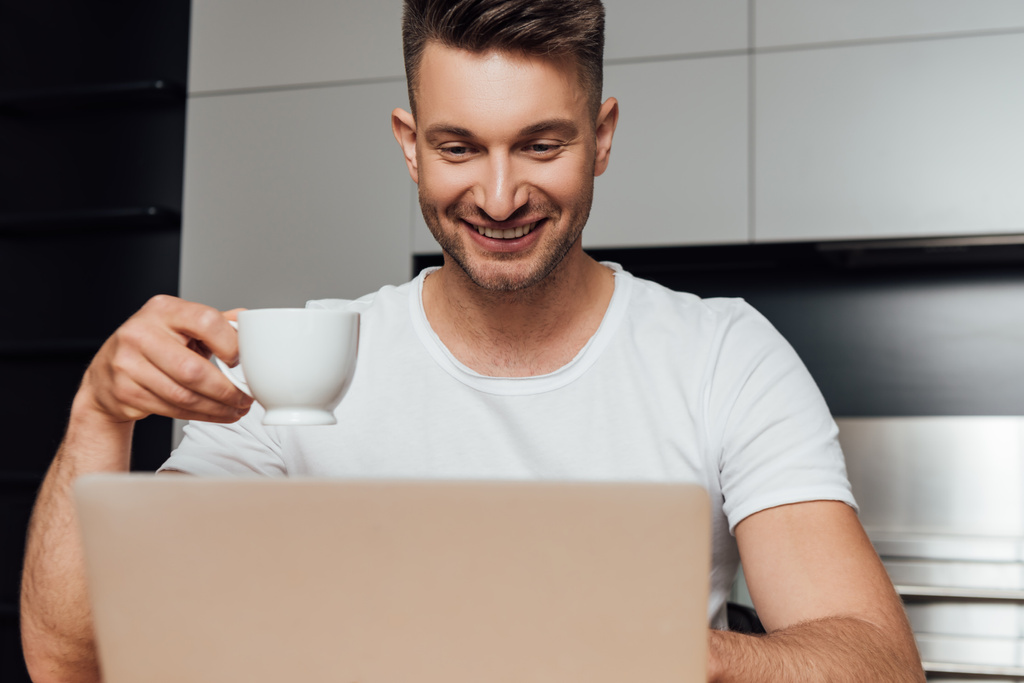 selective focus of happy freelancer holding cup of coffee and looking at laptop  - Photo, Image