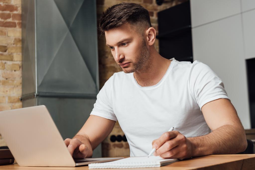 εστιασμένος άνθρωπος κρατώντας στυλό και κοιτάζοντας φορητό υπολογιστή, ενώ σε απευθείας σύνδεση μελέτη  - Φωτογραφία, εικόνα