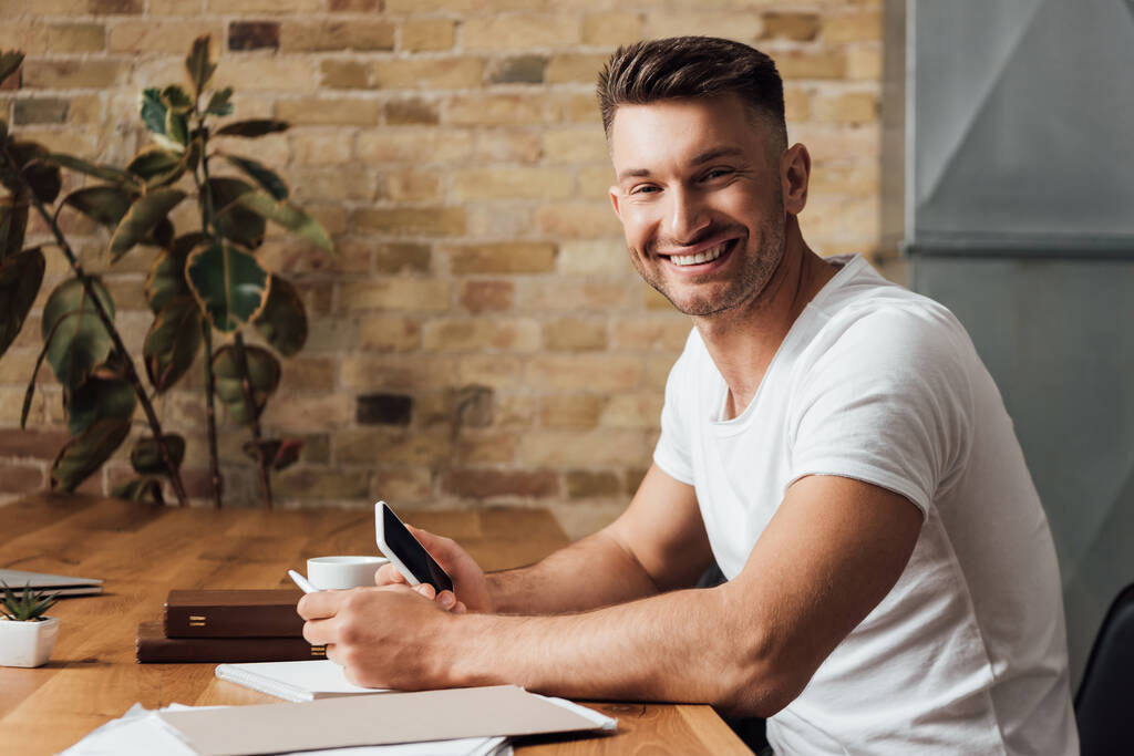 Πλευρική άποψη του ανθρώπου χαμογελώντας στην κάμερα, ενώ κρατώντας smartphone κοντά σε έγγραφα, βιβλία και κύπελλο στο τραπέζι  - Φωτογραφία, εικόνα