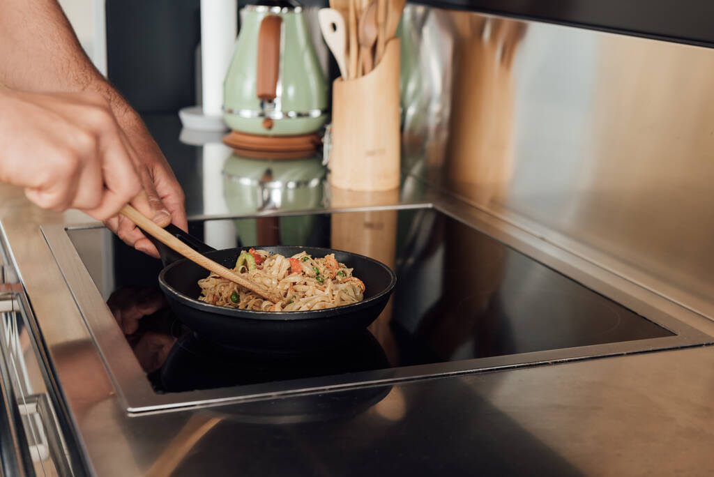 Gehakt uitzicht op de mens met houten spatel tijdens het bereiden van noedels in koekenpan in de keuken  - Foto, afbeelding