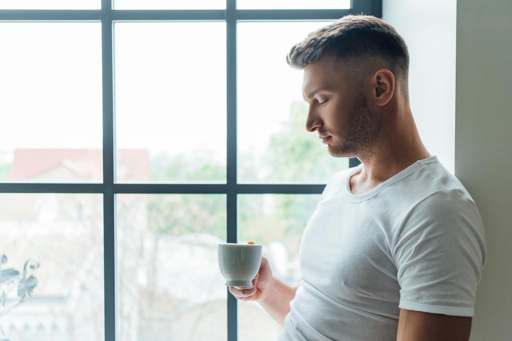 Πλευρική άποψη του όμορφου άνδρα κρατώντας φλιτζάνι καφέ, ενώ στέκεται κοντά στο παράθυρο στο σπίτι  - Φωτογραφία, εικόνα