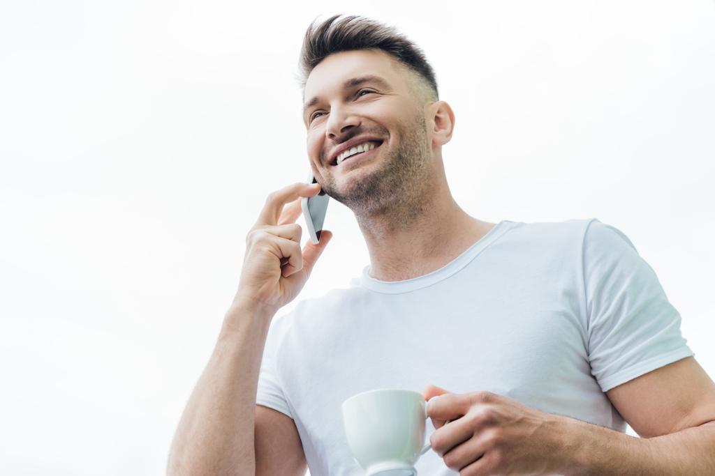Tiefansicht eines lächelnden Mannes, der auf dem Smartphone spricht und eine Tasse Kaffee isoliert auf weiß hält  - Foto, Bild