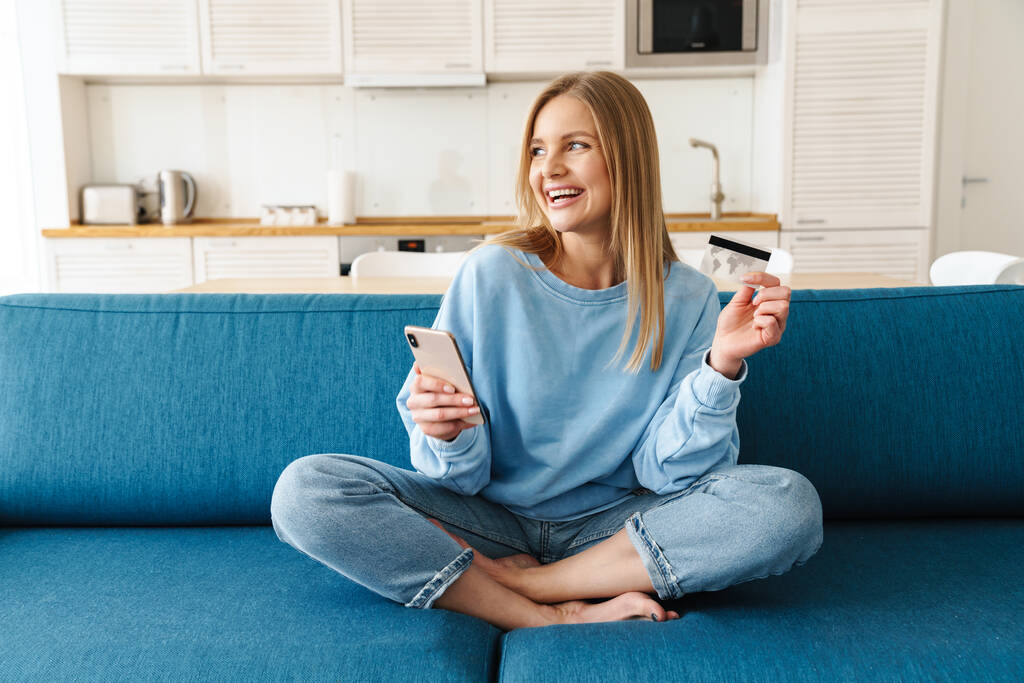 Immagine di una giovane donna sorridente che usa lo smartphone e tiene la carta di credito seduta sul divano di casa
 - Foto, immagini