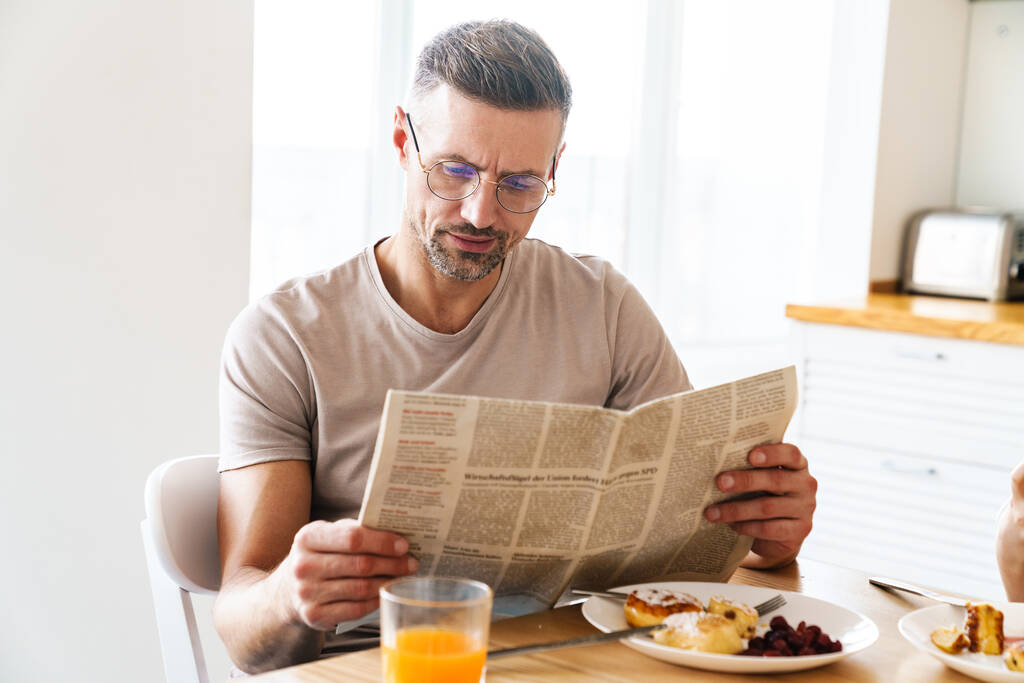 Fénykép komoly kaukázusi férfi szemüvegben újságot olvas miközben reggelizik a modern konyhában - Fotó, kép