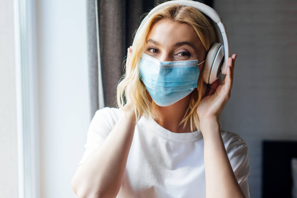 jovem mulher em máscara médica tocando fones de ouvido sem fio em casa
  - Foto, Imagem