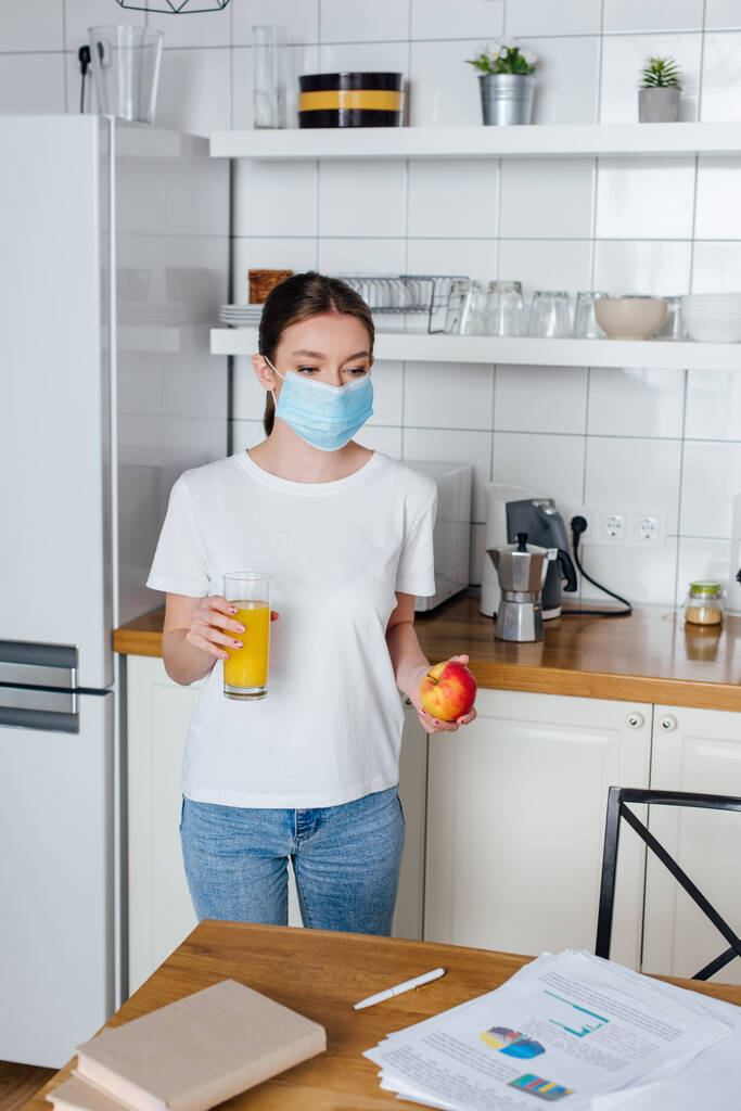 jonge vrouw in medisch masker met glas sinaasappelsap en appel - Foto, afbeelding