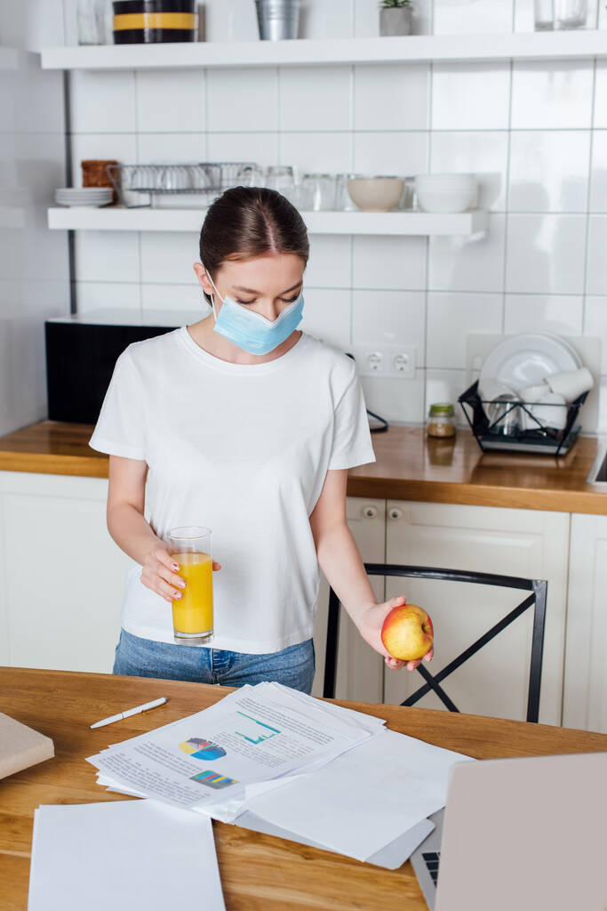 selective focus of young freelancer in medical mask holding glass of orange juice and apple near laptop - Photo, Image