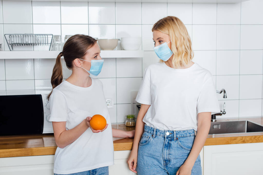 jovem mulher em máscara médica segurando laranja perto da irmã
 - Foto, Imagem