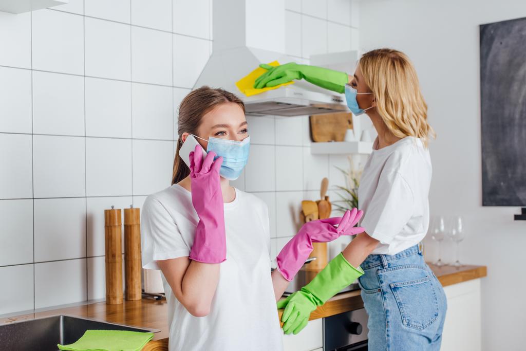 selektivní zaměření ženy v lékařské masce mluvení na smartphone v blízkosti sestra čištění kuchyně  - Fotografie, Obrázek